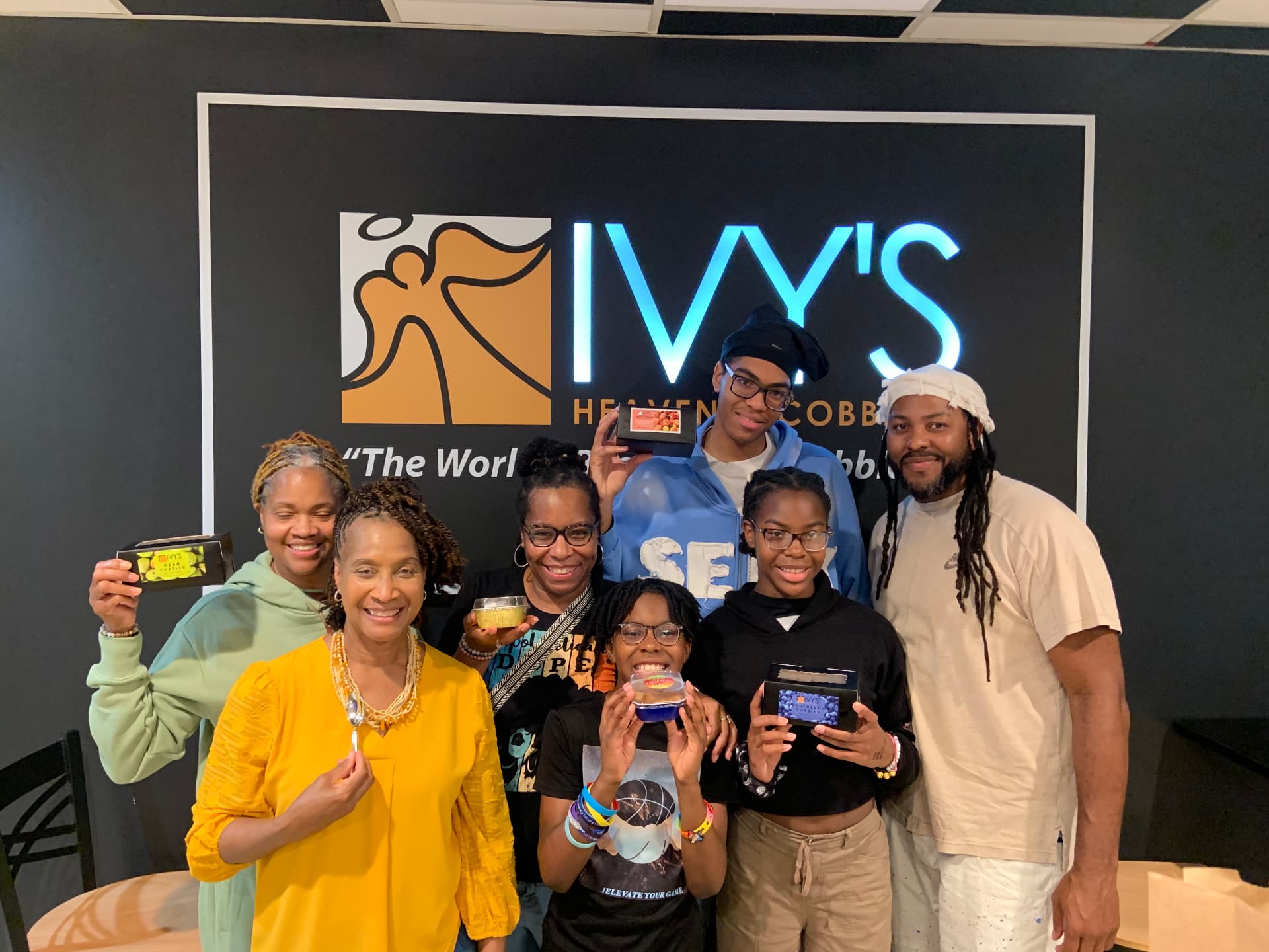 Family holding Ivy's cobblers in front of the lit Ivy's logo wall inside the shop